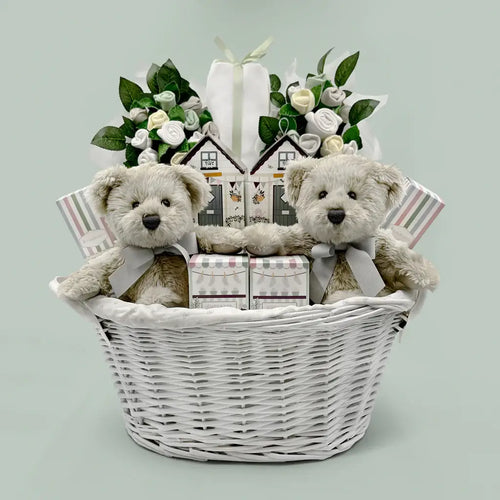 Luxury twins baby gift basket with white wicker basket holding two cream teddy bears with gray ribbons, two white rolled onesie bouquets with green leaves, double house-shaped gift box, and striped gift bags, sage green background