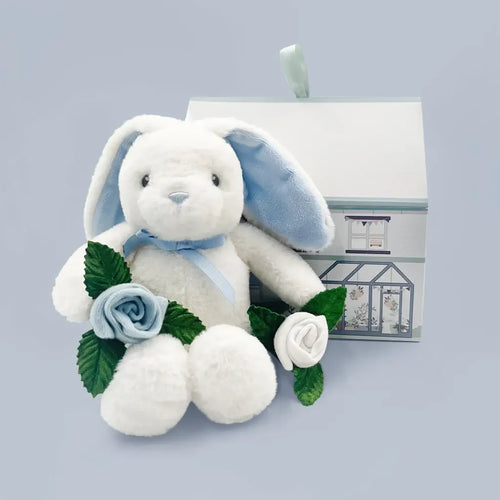 Luxury newborn gift box for baby boys with white plush bunny with blue ears and blue bow tie, one blue and one white rolled sock rosebud with green leaves, illustrated greenhouse house-shaped gift box, blue-gray background