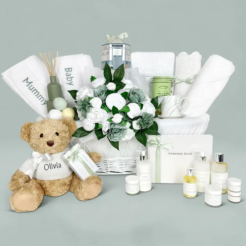 Luxury newborn gift collection in sage green and white: personalized teddy bear in knitted sweater, rolled baby clothes bouquet, embroidered Mommy and Baby towels, keepsake book, skincare bottles, candle, reed diffuser, fine china mug, and biscuit tin, sage green background.