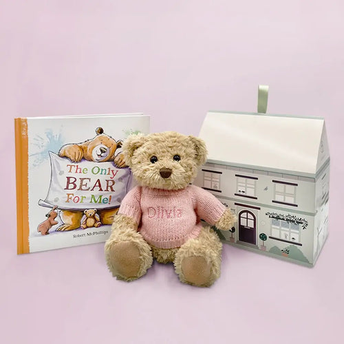 Tan plush teddy bear in a pink embroidered knit sweater with the name "Olivia" beside "The Only Bear For Me!" hardcover picture book and a house-shaped illustrated gift box — personalized sibling bear and book gift set.
