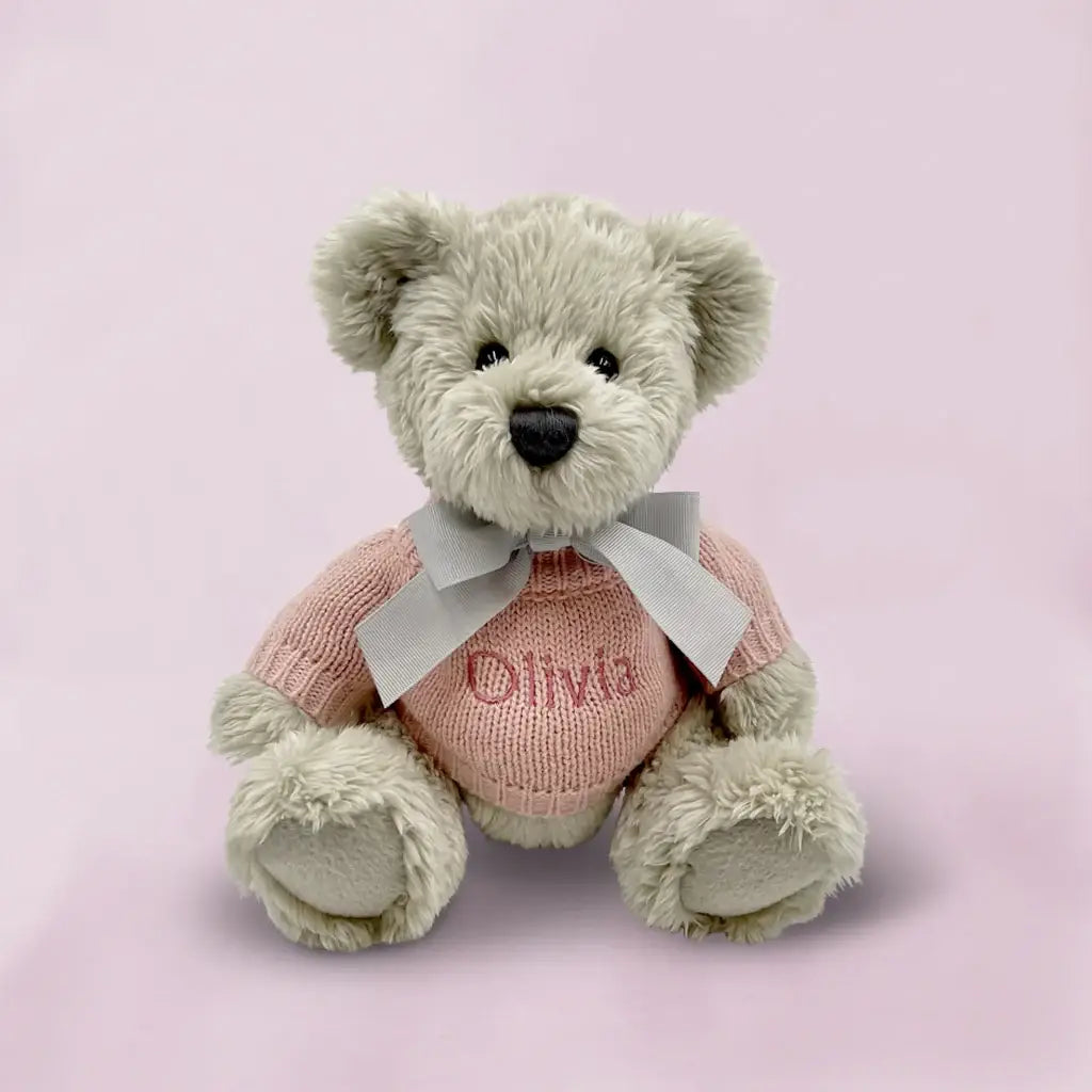 Personalized grey teddy bear wearing a pink knitted sweater embroidered with a baby girl's name, presented in an illustrated gift box
