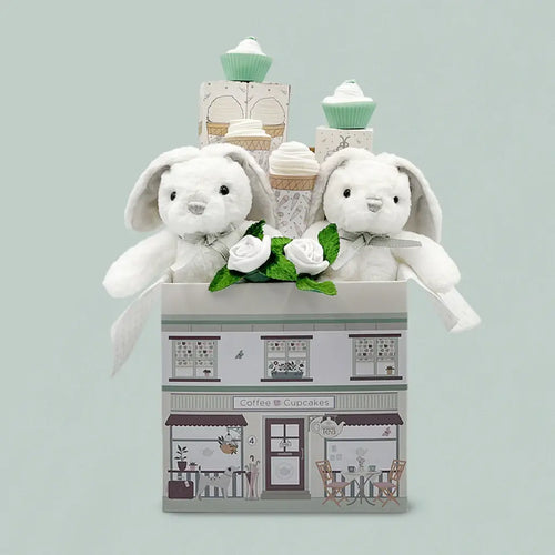 Twins newborn gift box with two white plush gray-eared bunnies with white ribbon bows, white rolled sock rosebuds with green leaves, two mint cupcake sock towers, illustrated coffee and cupcakes house-shaped gift box, sage green background