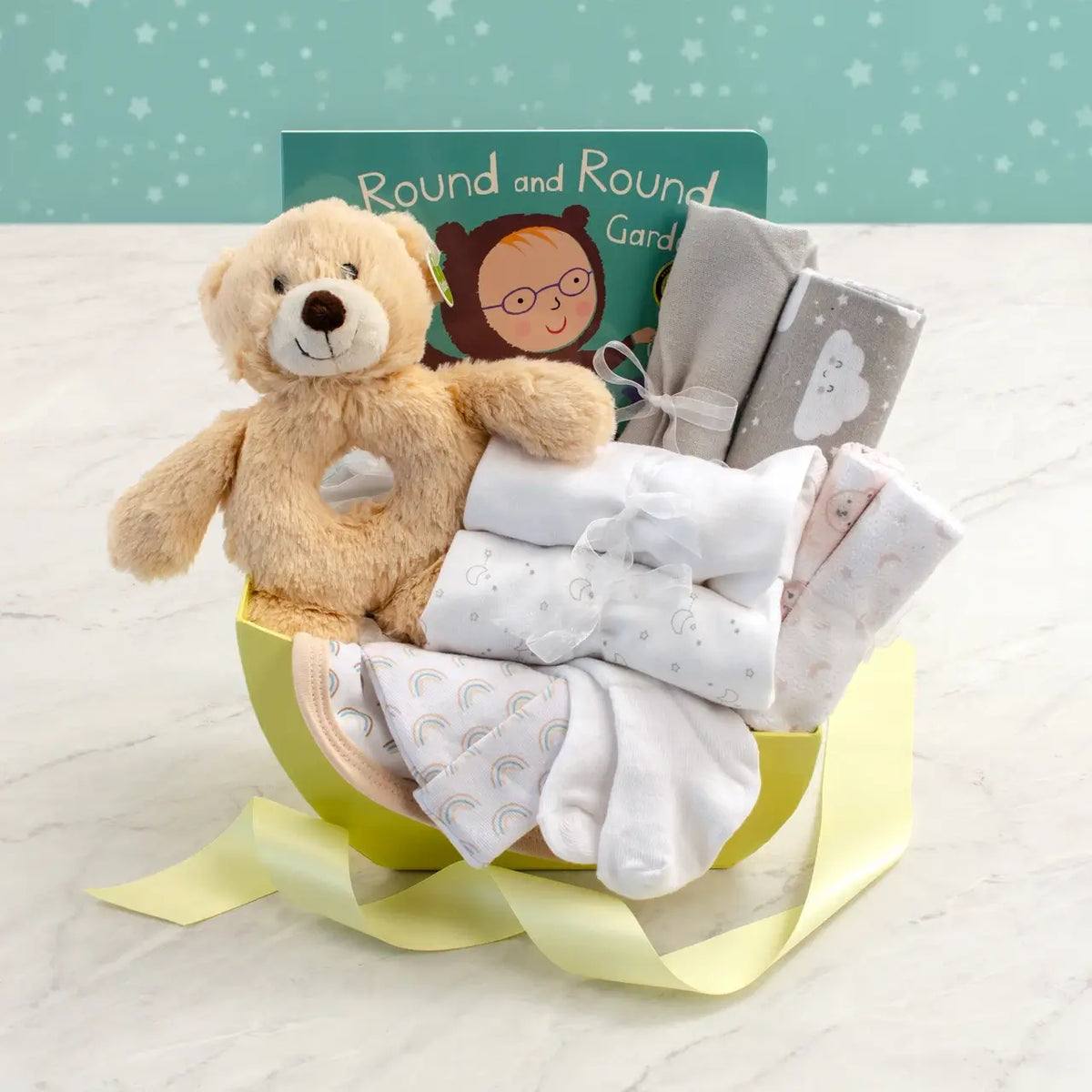 Gender-neutral newborn gift basket with plush teddy bear rattle, board book, and soft layette essentials in cradle-shaped yellow basket