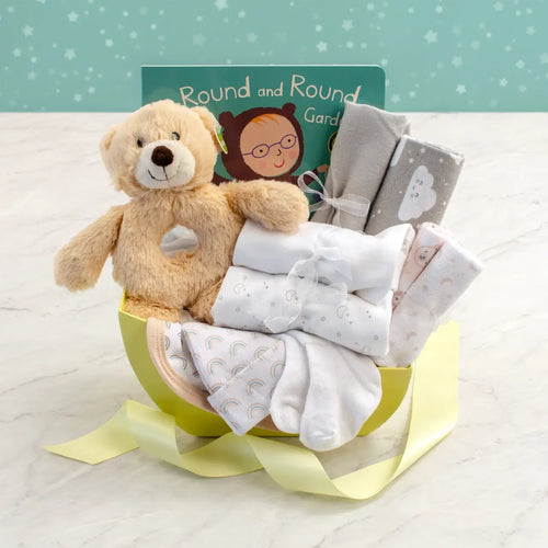 Gender-neutral newborn gift basket with plush teddy bear rattle, board book, and soft layette essentials in cradle-shaped yellow basket