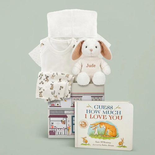 Personalized beige bunny with embroidered name on knitted sweater, white baby bathrobe, bunny-print pajamas, muslin swaddle, and Guess How Much I Love You board book in illustrated gift box, sage green background.