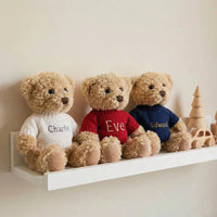 Three personalized first Christmas teddy bears in red, navy, and white sweaters on a nursery wall shelf