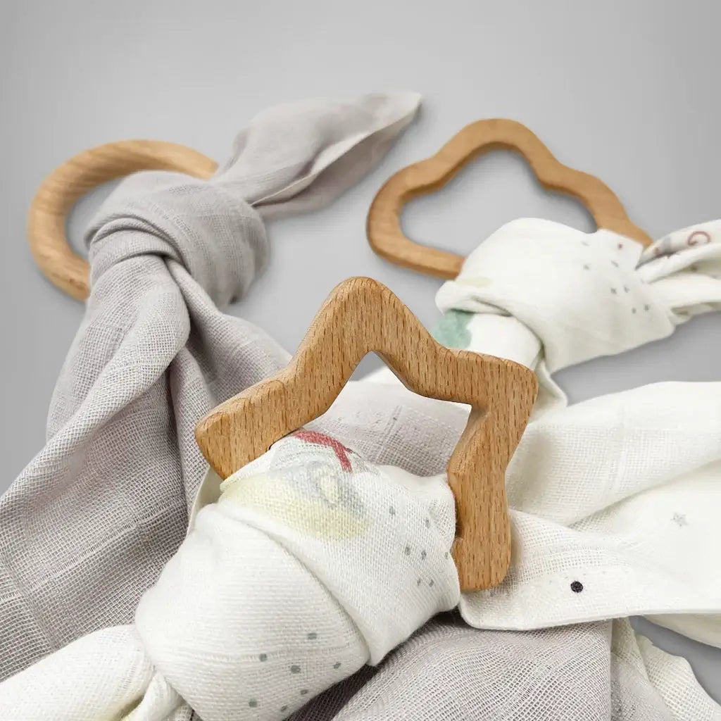 Three wooden baby teethers in star, circle, and cloud shapes each tied with a soft muslin cloth