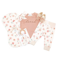7-piece organic baby girl layette set flat lay — mauve floral bodysuit, leggings, booties, headband, teether, and burp cloth