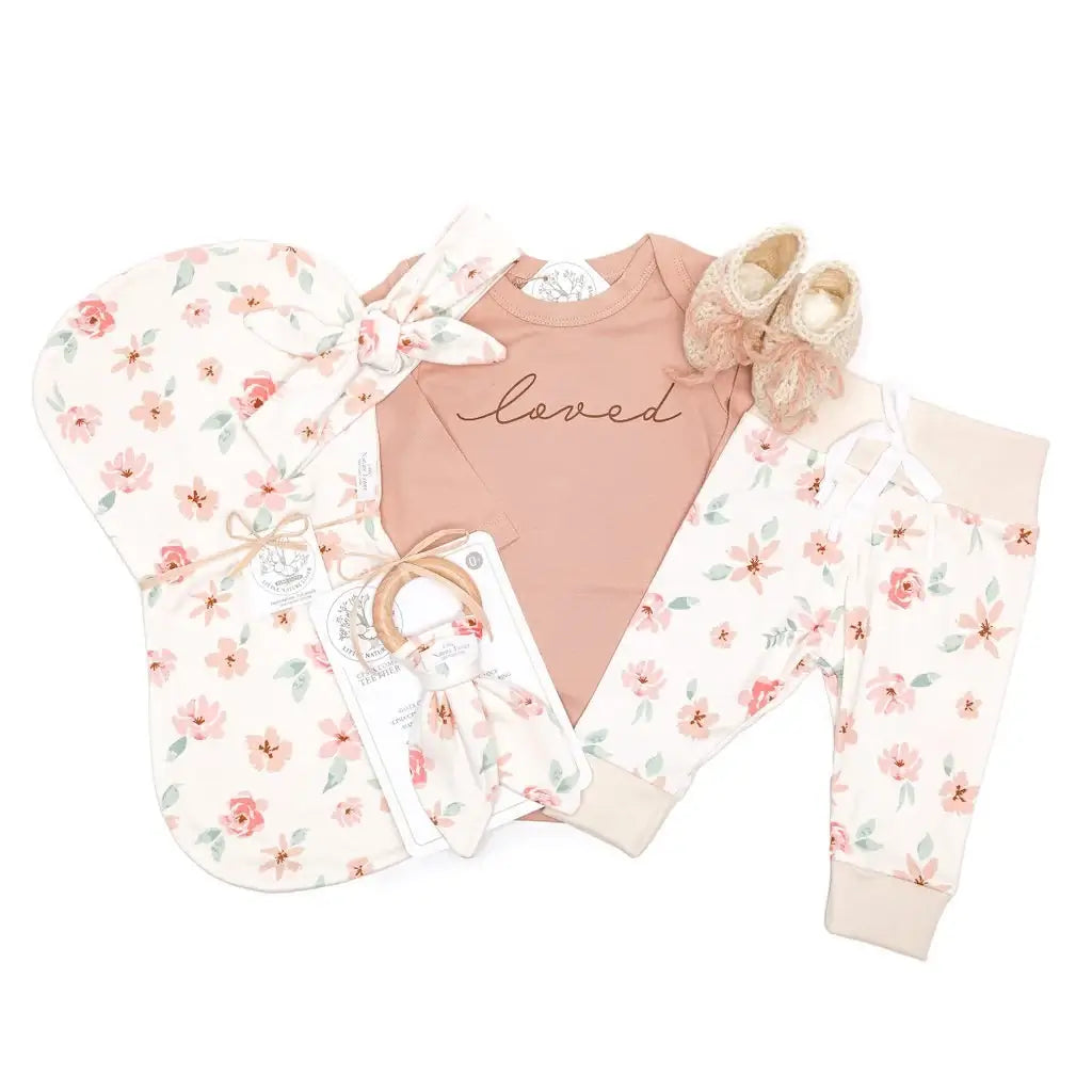 7-piece organic baby girl layette set flat lay — mauve floral bodysuit, leggings, booties, headband, teether, and burp cloth