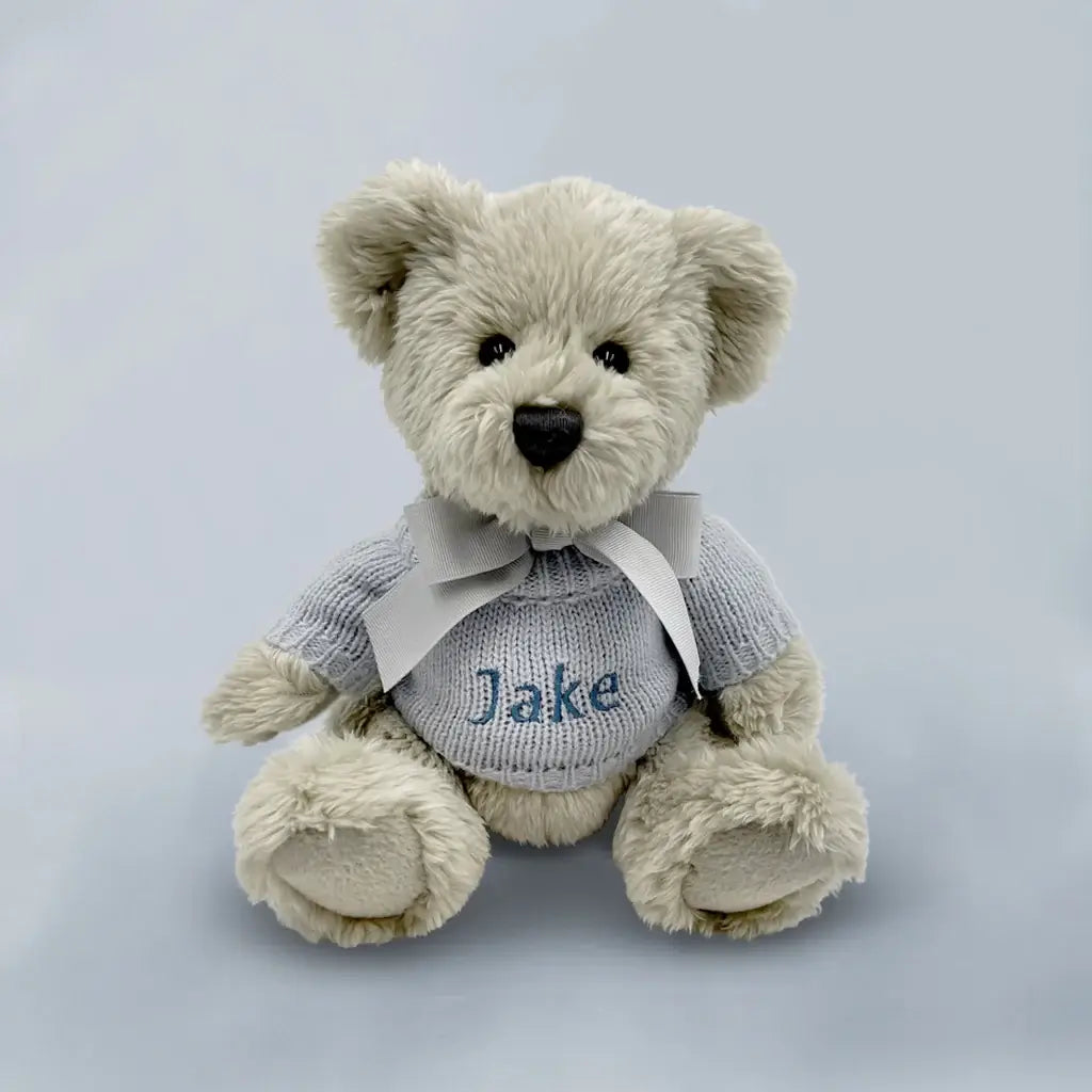 Personalized grey teddy bear wearing a blue knitted sweater embroidered with a baby boy's name, presented in an illustrated gift box