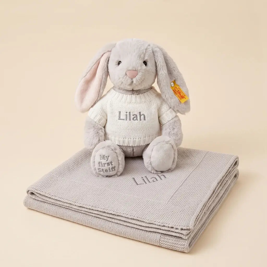 Personalized My First Steiff Hoppie Rabbit in grey with embroidered name on knitted sweater, paired with a personalized cotton knitted blanket in illustrated gift box