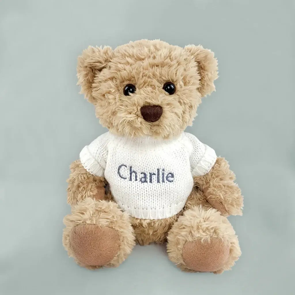Personalized grey teddy bear wearing a white knitted sweater embroidered with a baby's name, presented in an illustrated gift box